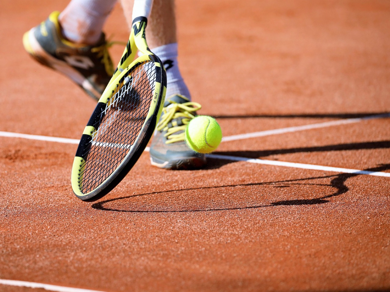 Raqueta y pelota sobre una pista de tierra batida junto a la línea, con las zapatillas de un jugador